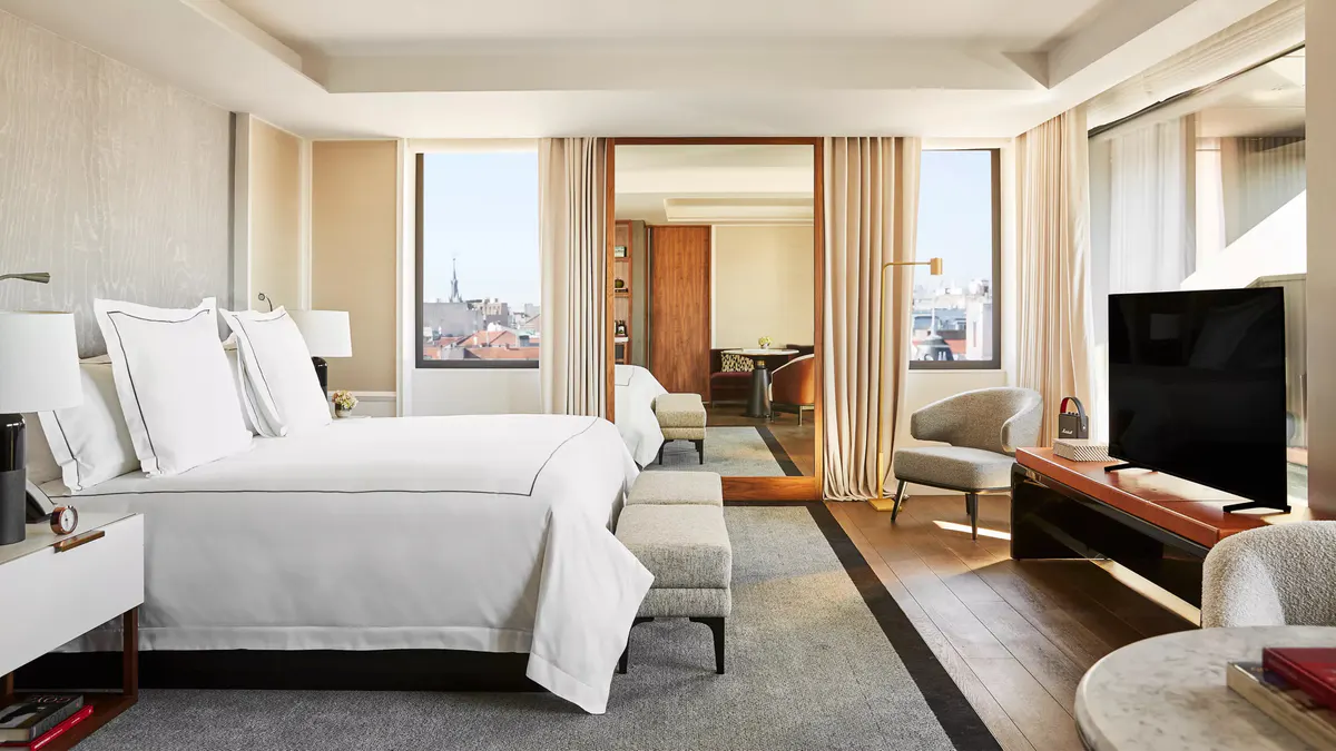 A guest room with white bedding, floor-to-ceiling windows with city views, and warm wood accents