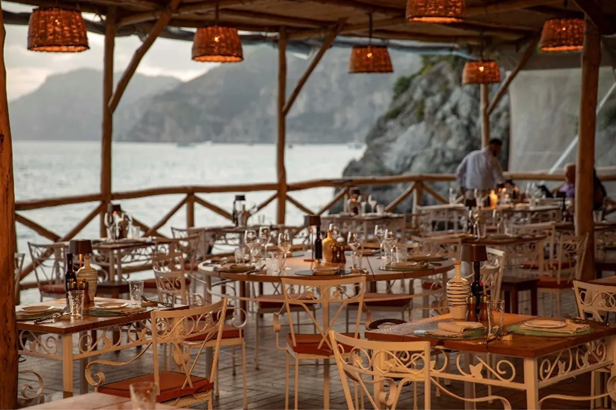 Covered restaurant interior with wooden chairs, round tables, white linens, and Mediterranean coastline backdrop
