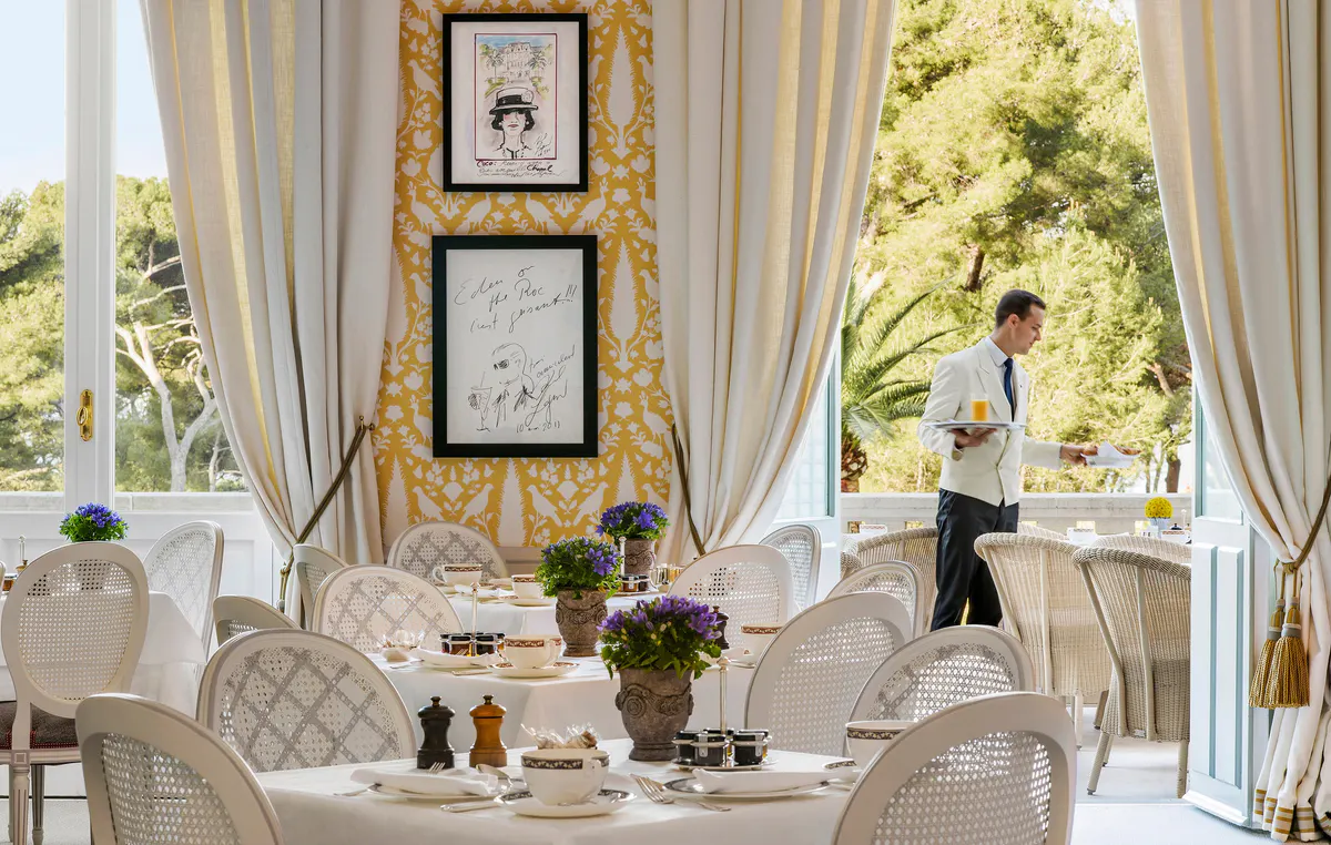 An elegant dining room with cream drapery, framed artwork, white tablecloths, and garden views