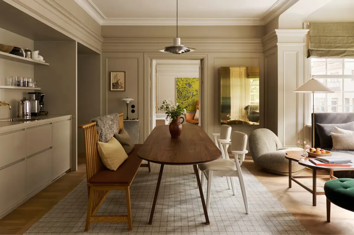 A suite with dining table, wooden chairs, bookshelves, and artwork on taupe walls with bright windows