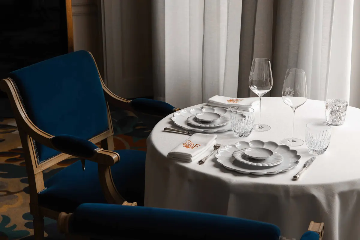 A place setting with white linens, polished flatware, and blue upholstered chair