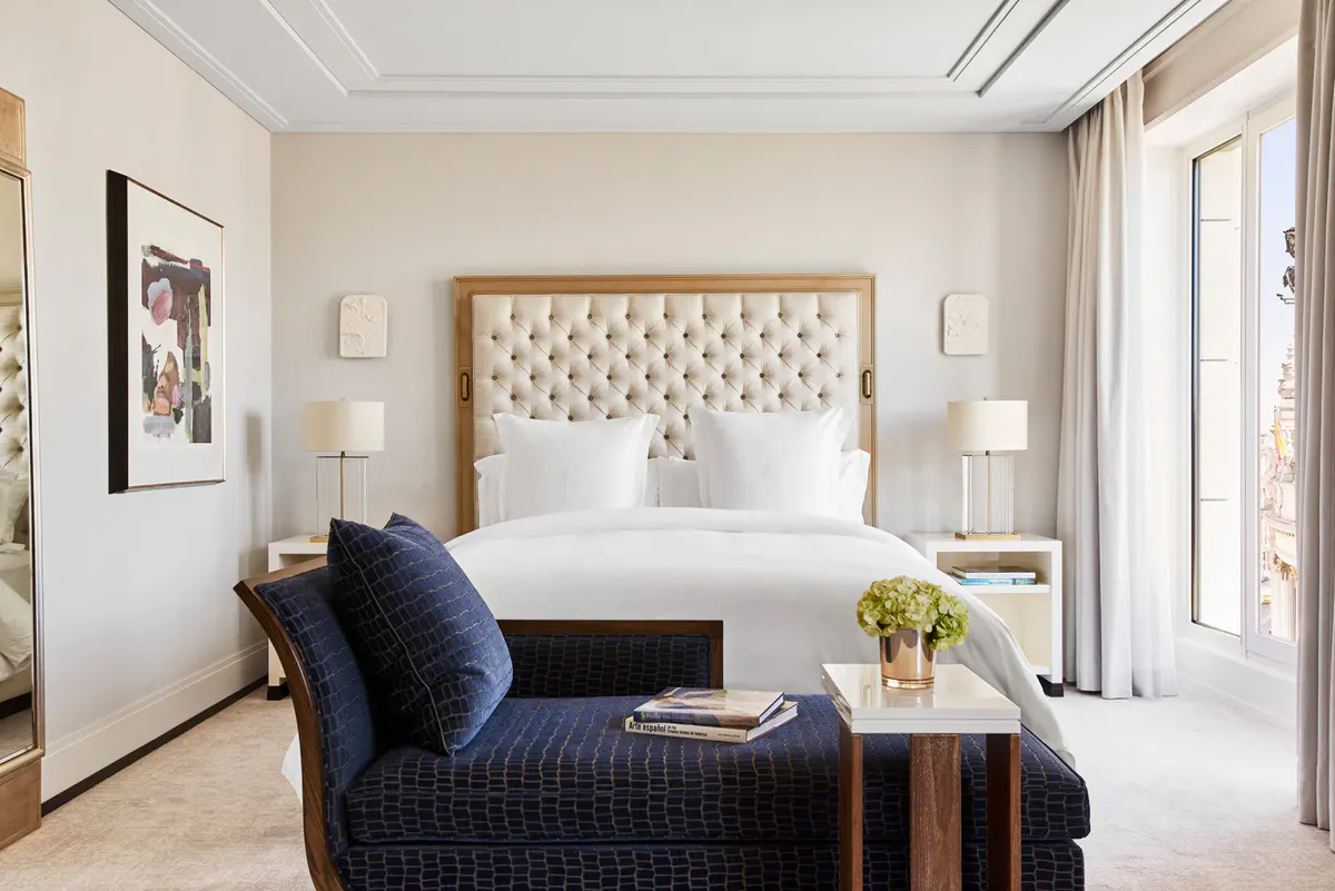 Suite bedroom with quilted white headboard, blue accent chair, and French doors with cream curtains