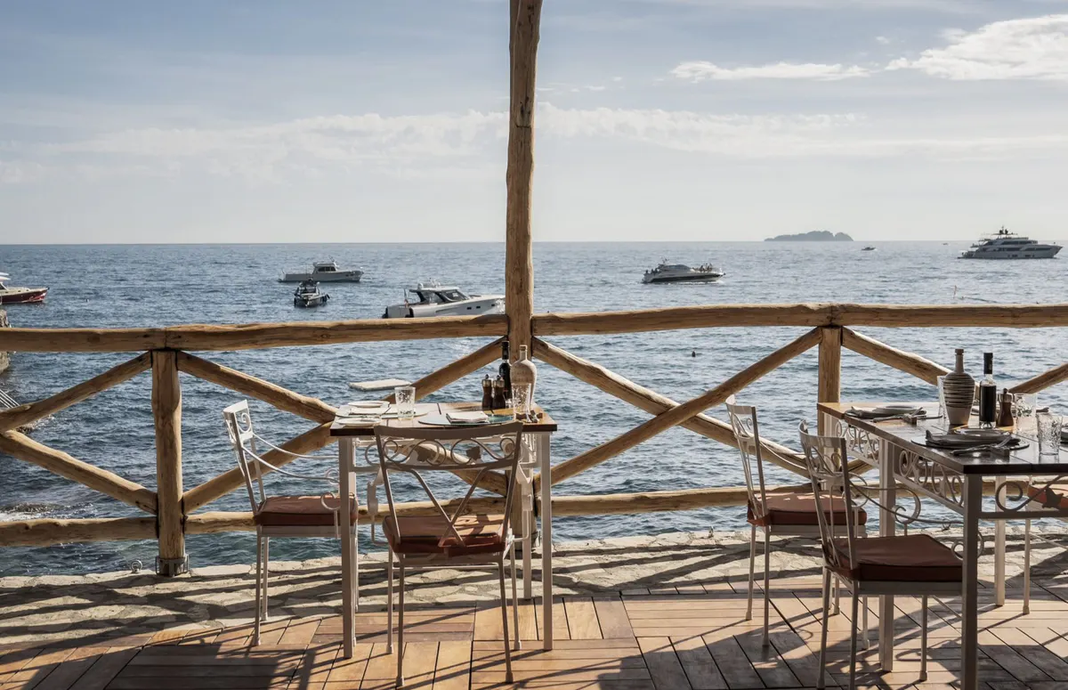 Seaside terrace with white-clothed tables, wooden chairs, and clear Mediterranean water view