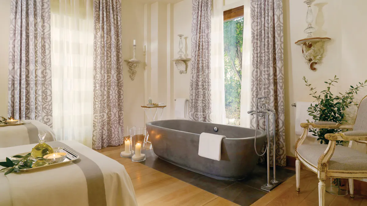 Spa bathroom with dark stone tub, tall windows with patterned drapery, and pale walls