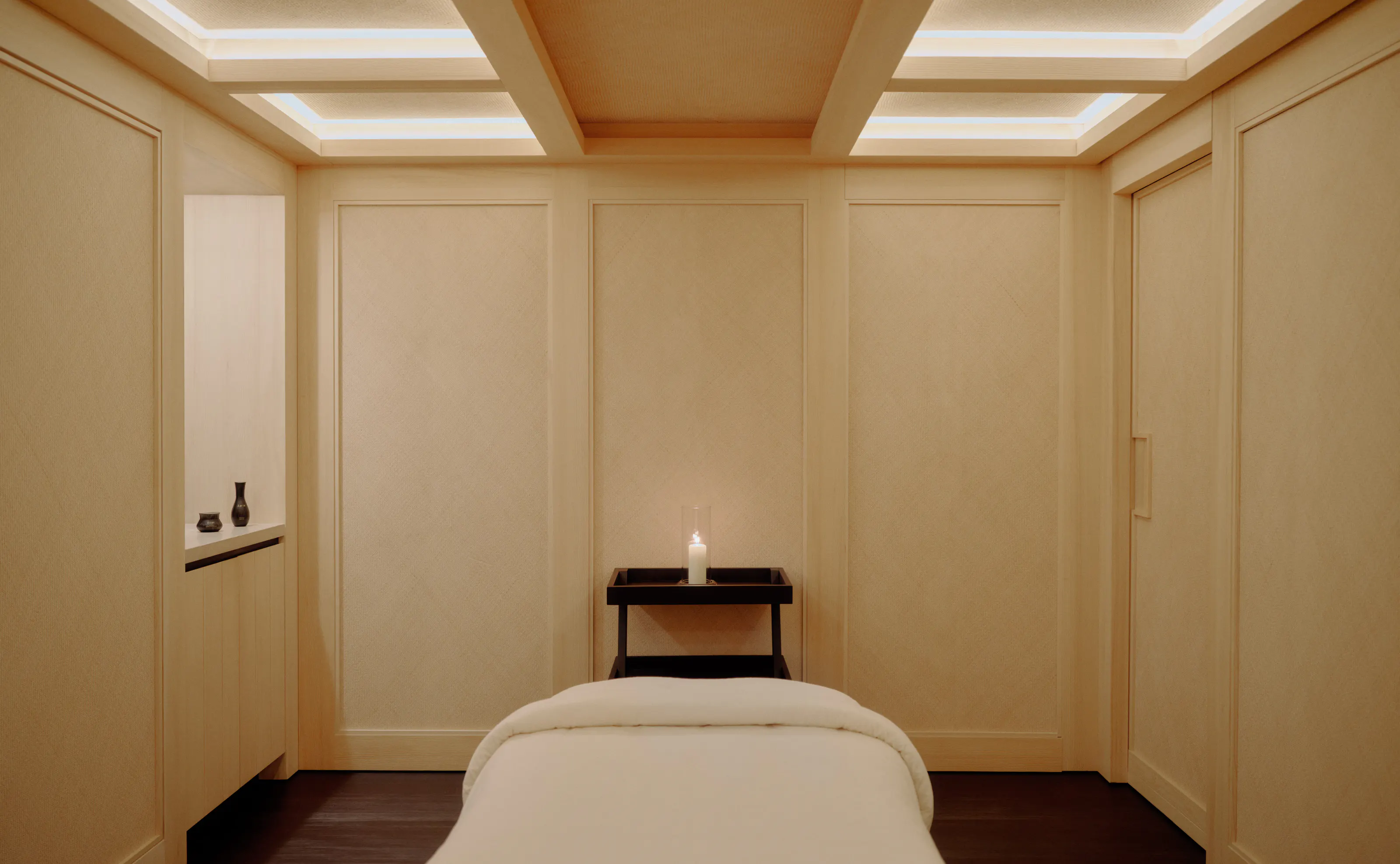 A serene Aman treatment room in pale oak, a candle on a low table below a backlit coffered ceiling