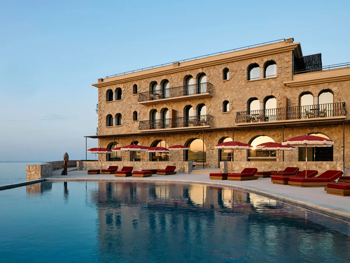 Fringed red parasols and loungers line the pool as the stone hotel glows at dusk