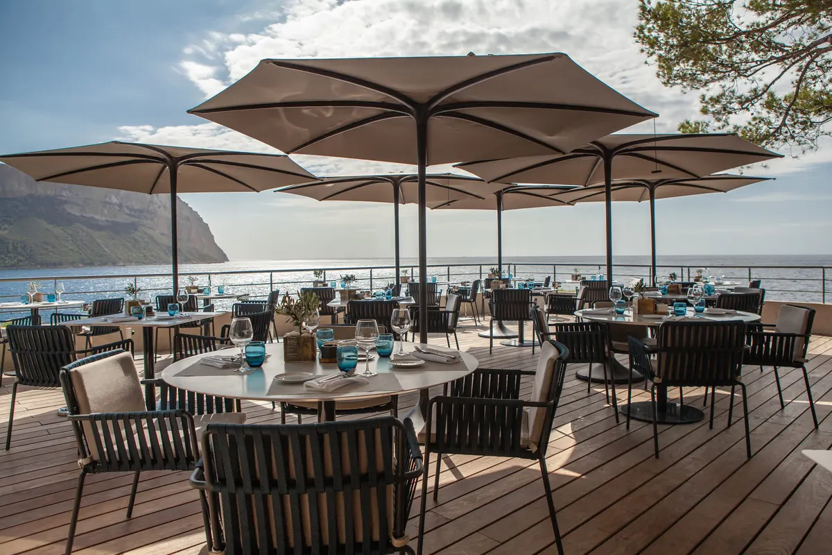 A seaside dining terrace with large umbrellas, wooden tables, and Mediterranean Sea beyond