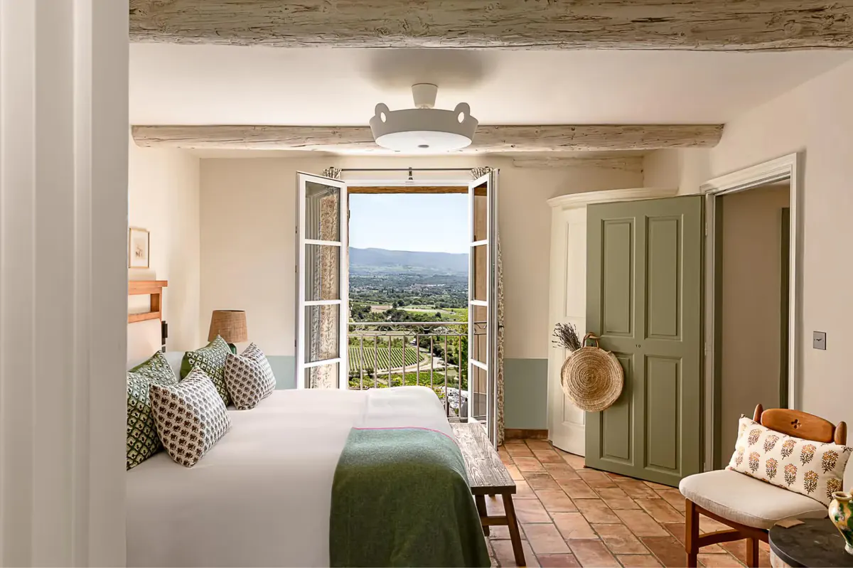 A bedroom with exposed wooden beams, green shutters, and an open door revealing a countryside vista