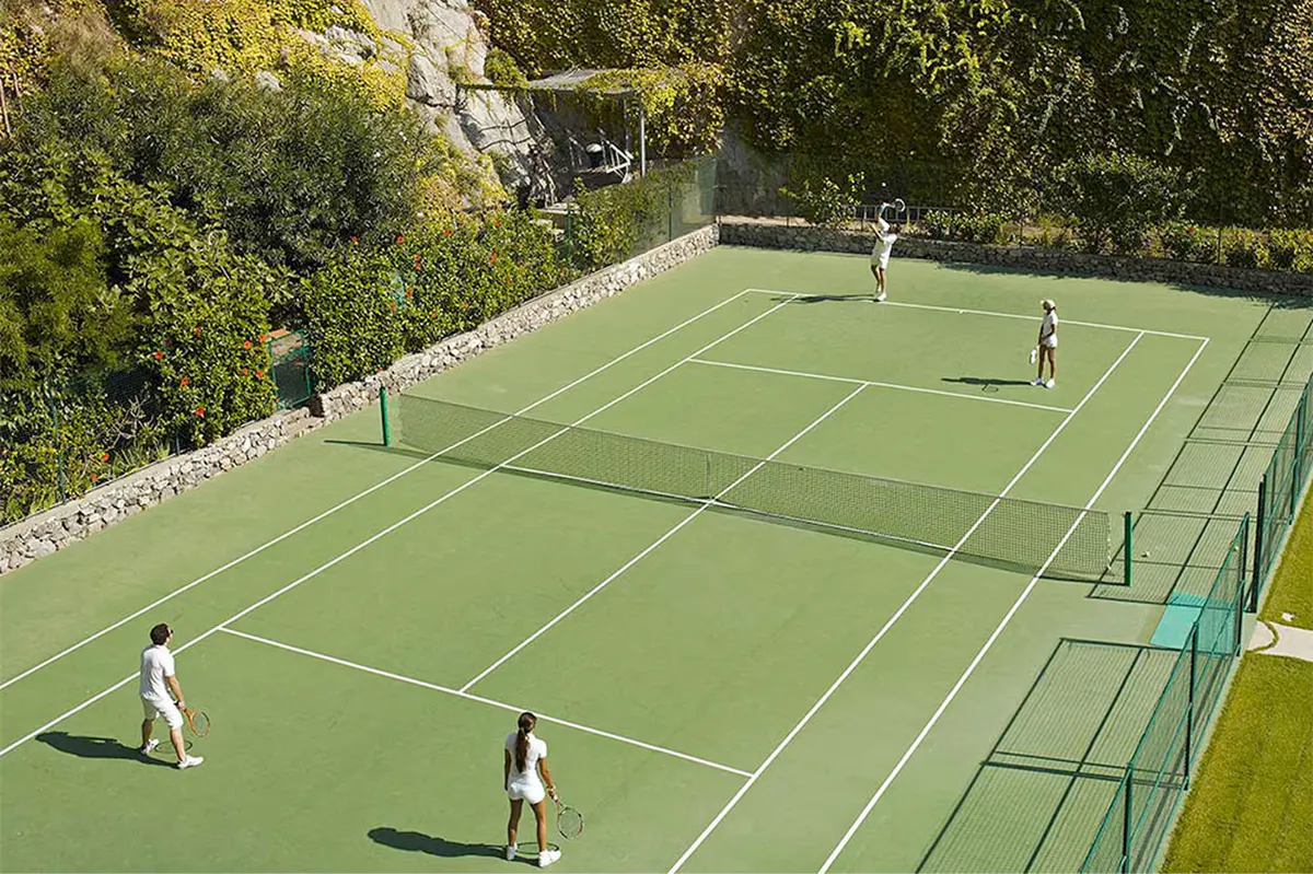 Tennis court at cliff edge with players, net, and panoramic sea and coastline views