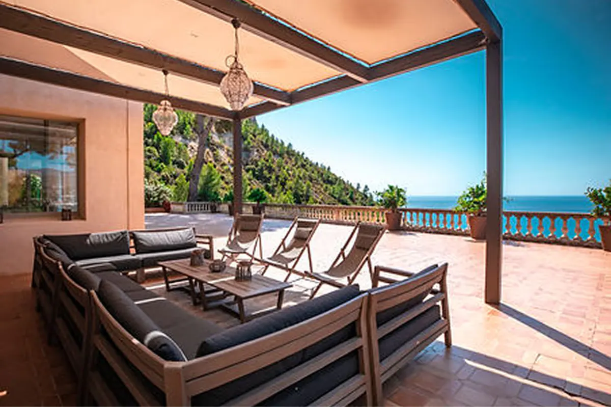 Covered terrace with dark wooden furniture and cushions overlooking turquoise sea and pines