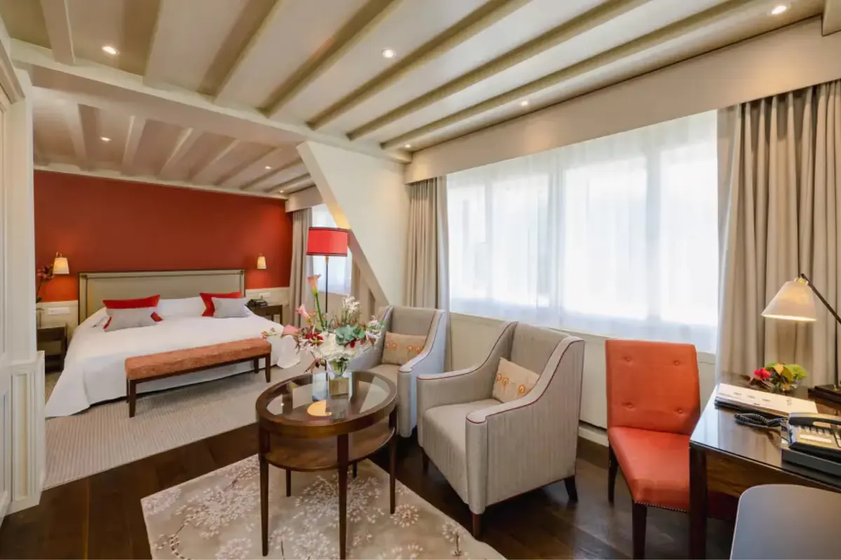 A bedroom with red headboard, lounge seating, wood beams, and bay windows with mountain views
