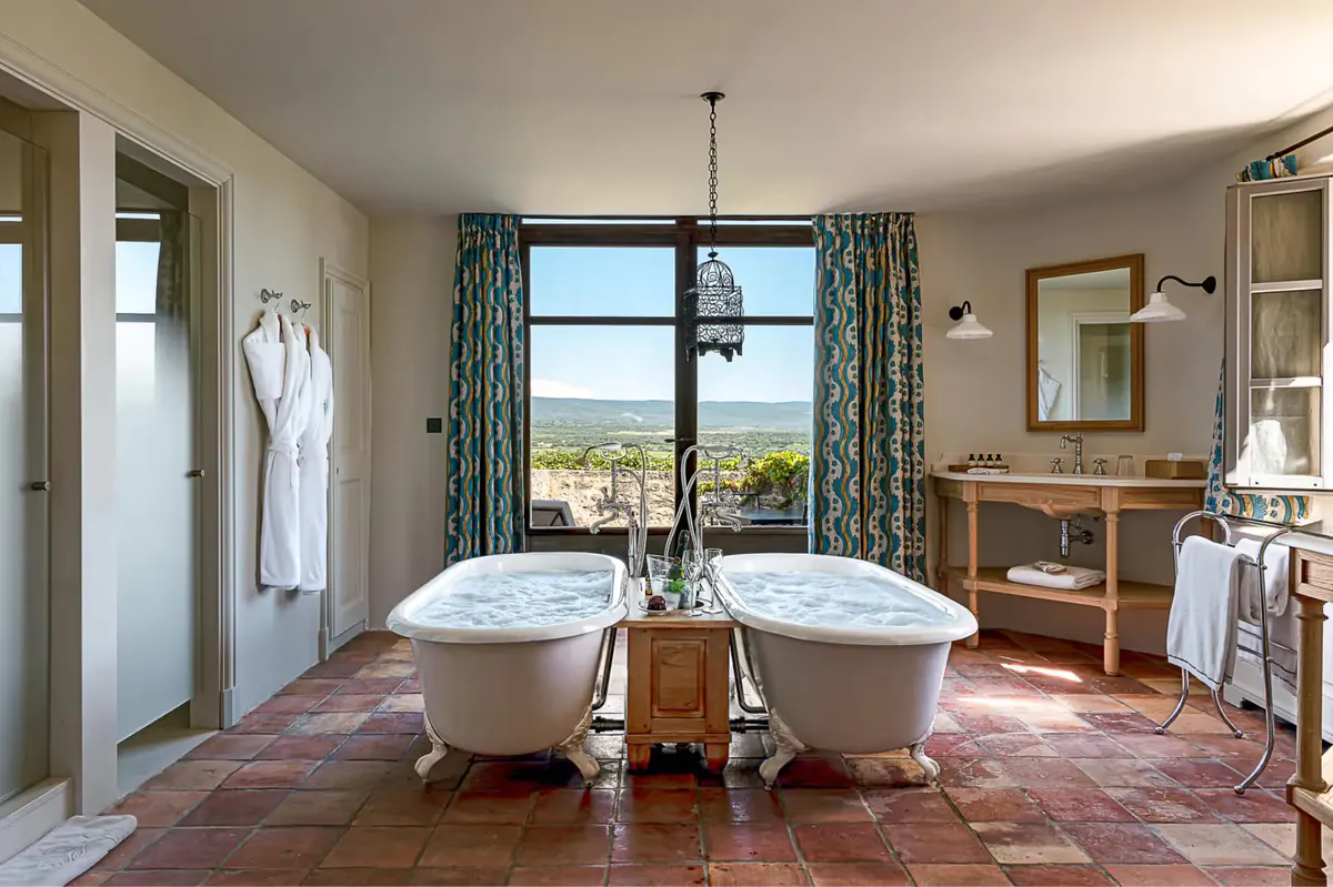 A spa-like bathroom with two white bathtubs, terracotta tile floor, and countryside views through large windows