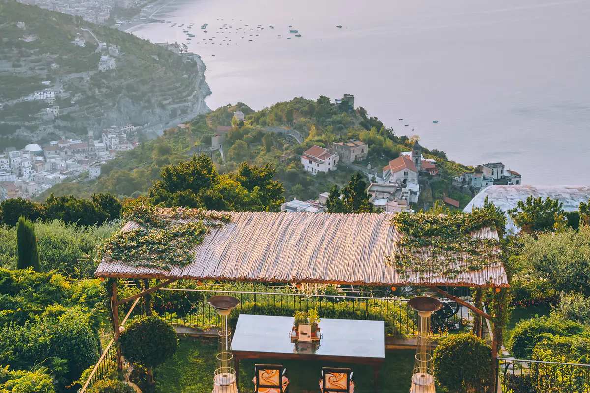 Aerial view of wooden pavilion with coastal dining setup on terraced gardens overlooking sea and village