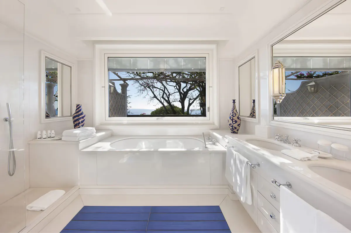 White marble bathroom with view window overlooking Mediterranean landscape, soaking tub, blue accent mat