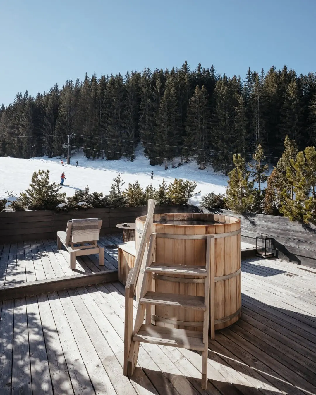 Wood barrel hot tub steaming on a sunlit deck above the piste and snow laden pines