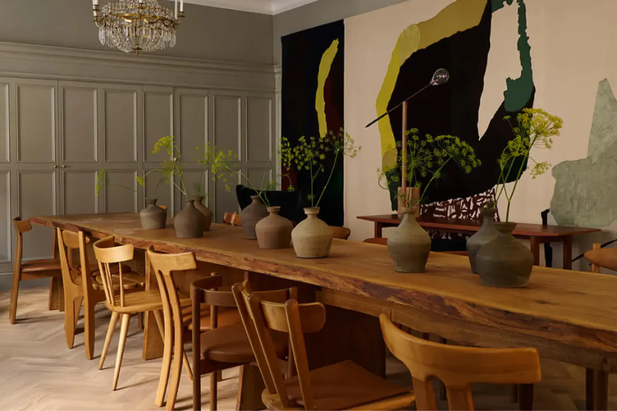 A dining room with wooden table and chairs, potted plants, and dark artwork on neutral walls