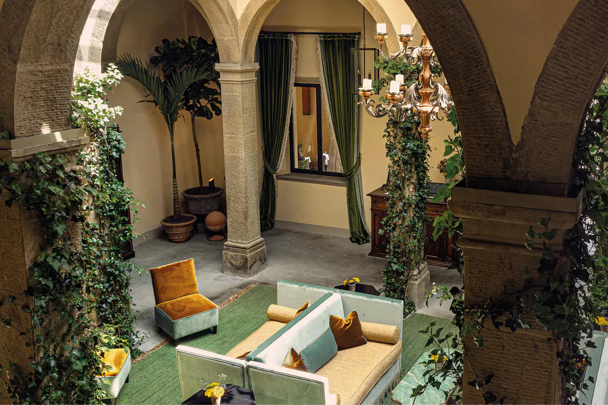 Monastic courtyard with barrel vaults, cream seating, green drapes, climbing ivy, and ornate gilt chandelier