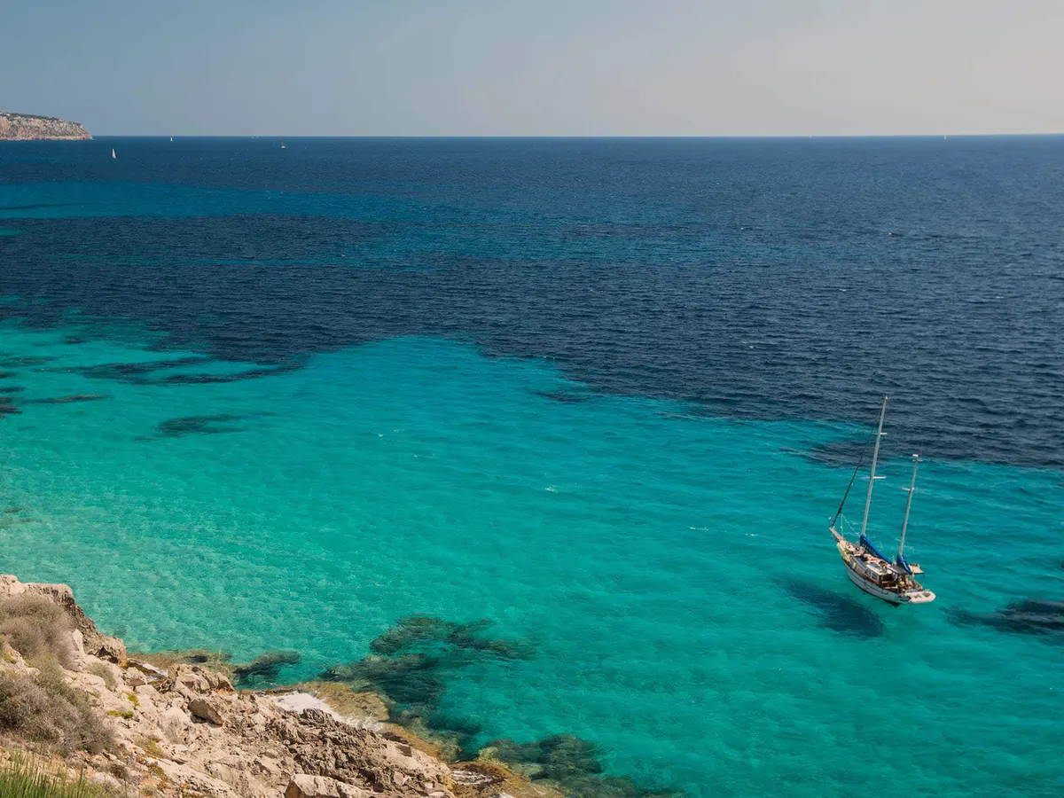 Turquoise coves and pale limestone cliffs frame a moored sailboat in clear waters
