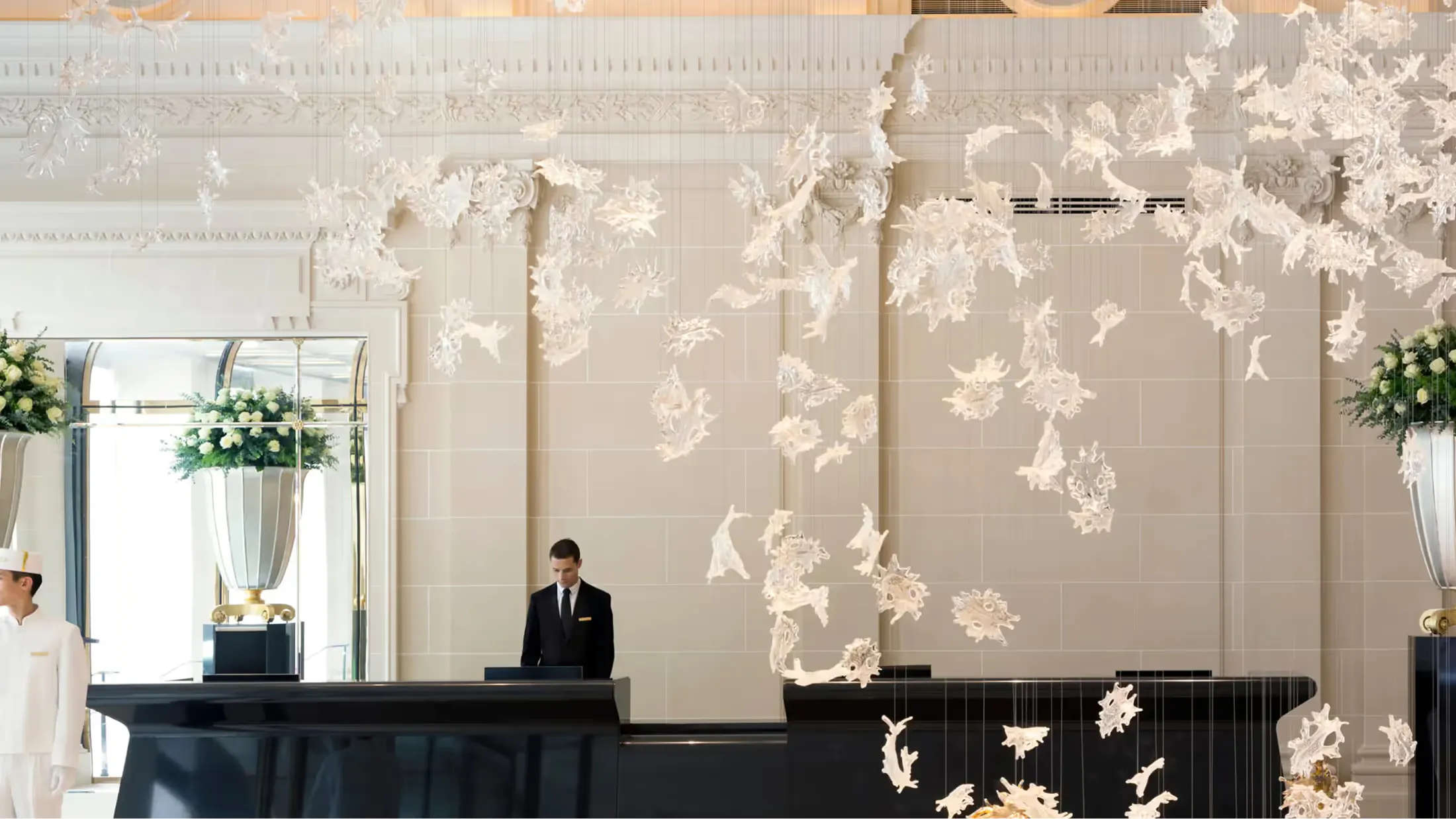 A refined Parisian lobby with cream-colored walls, suspended floral installations, and a concierge at a dark desk