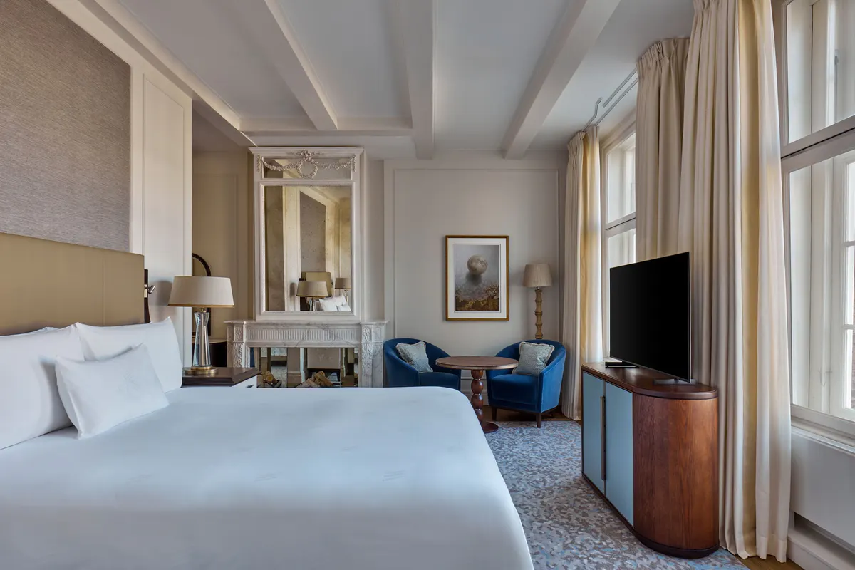 A guest room with a cream headboard, white bedding, blue accent chairs, and tall windows with draped curtains