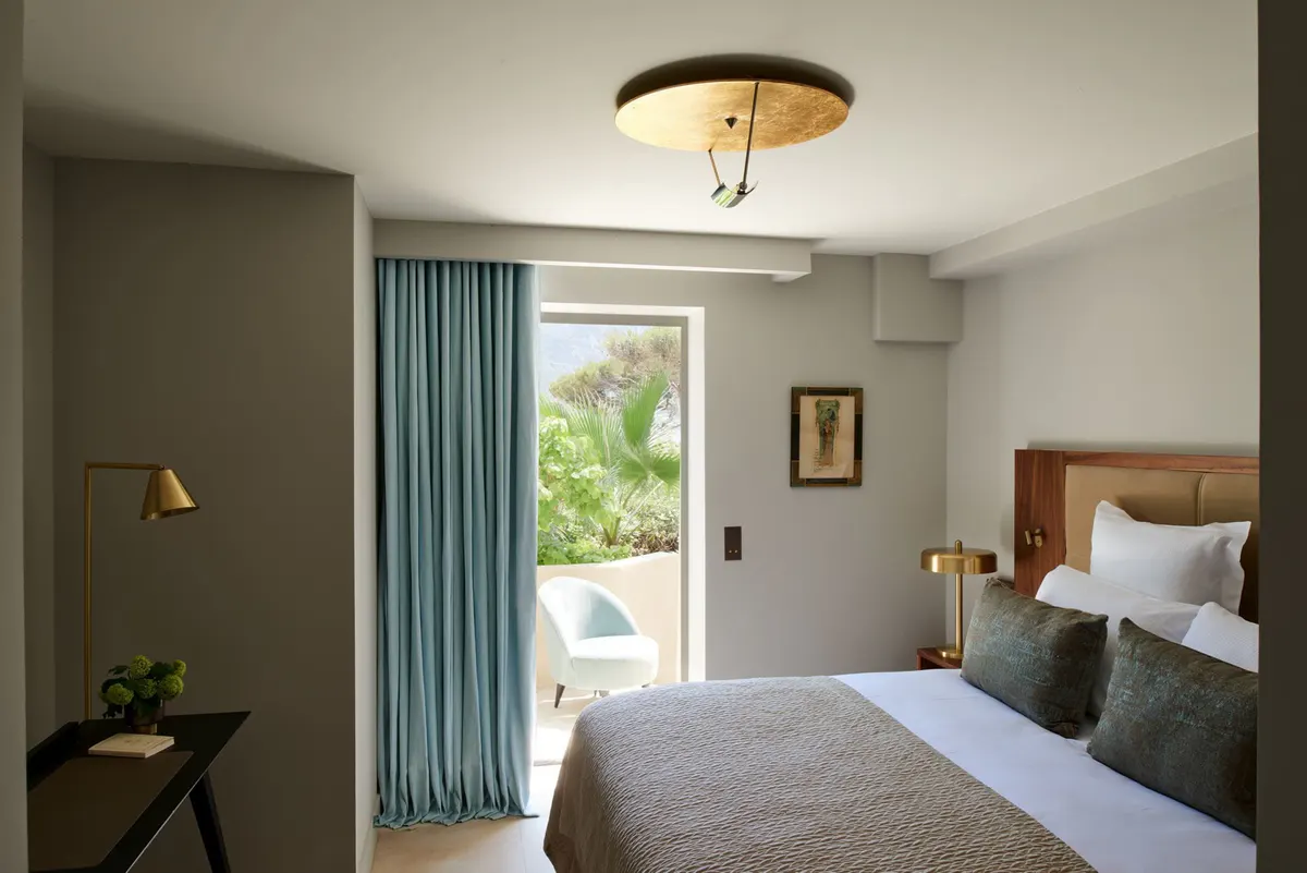 A bedroom with pale walls, brass ceiling fixture, and window with Mediterranean light