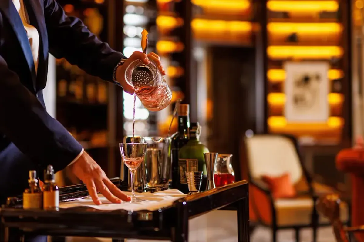 A hand pouring amber spirit into a glass at a marble bar counter with bottles in the background