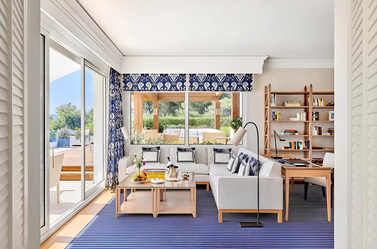 A sitting room with blue carpet, linen furniture, arched windows opening to sunlit grounds