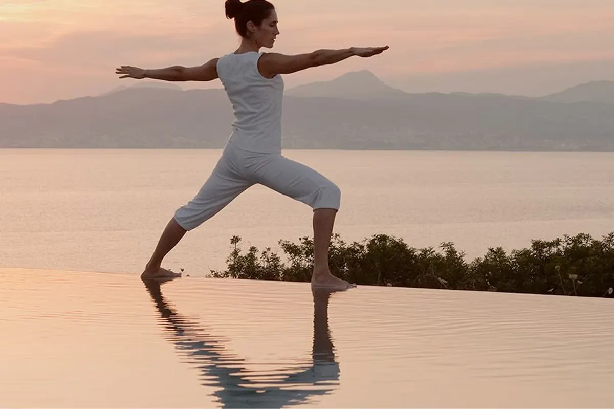 A figure practices yoga in a wide stance pose beside a reflecting pool at golden hour