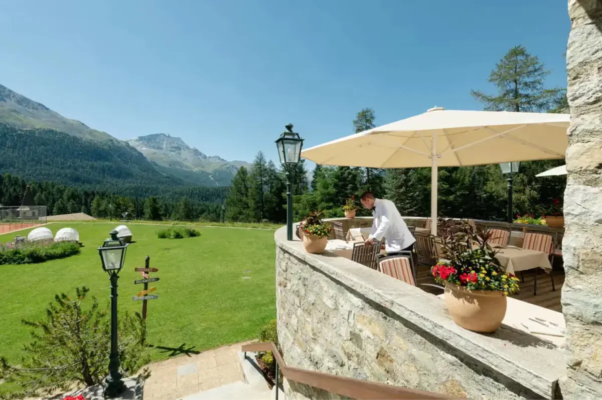 An outdoor terrace dining area with cream umbrella, verdant lawn, and mountain backdrop