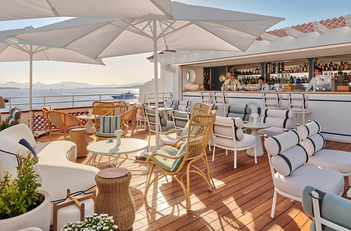 A sun-drenched terrace with white umbrellas, wooden deck, bar area, and Mediterranean panorama