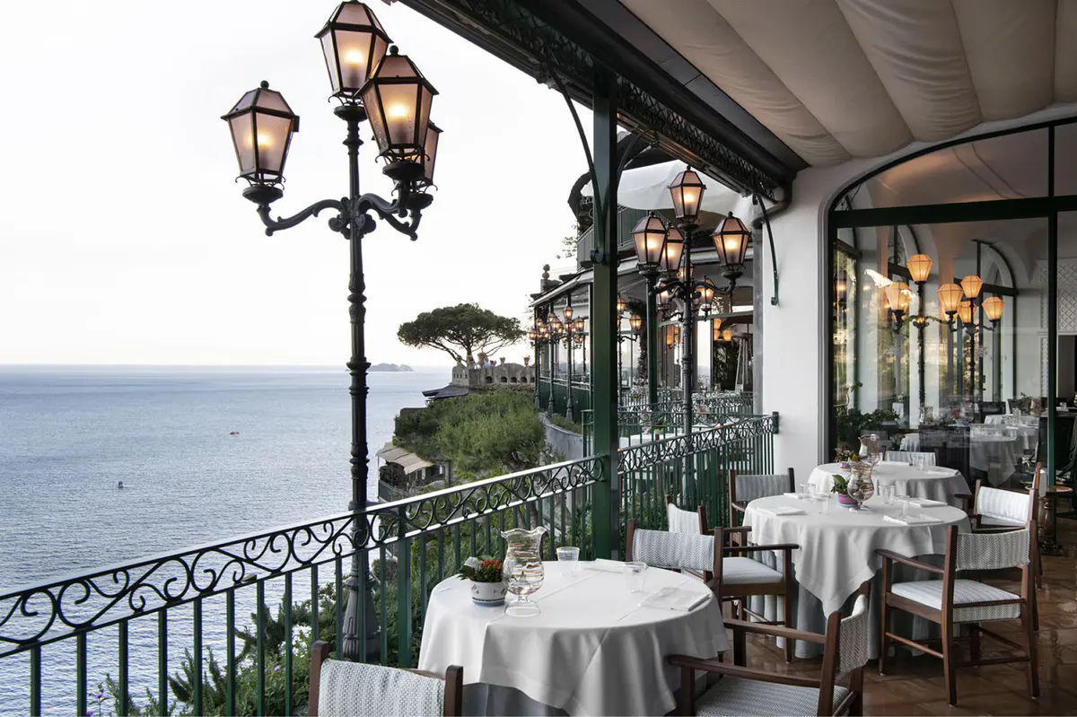 Elegant covered patio dining with white tablecloths, brass lanterns, railing, and sea views