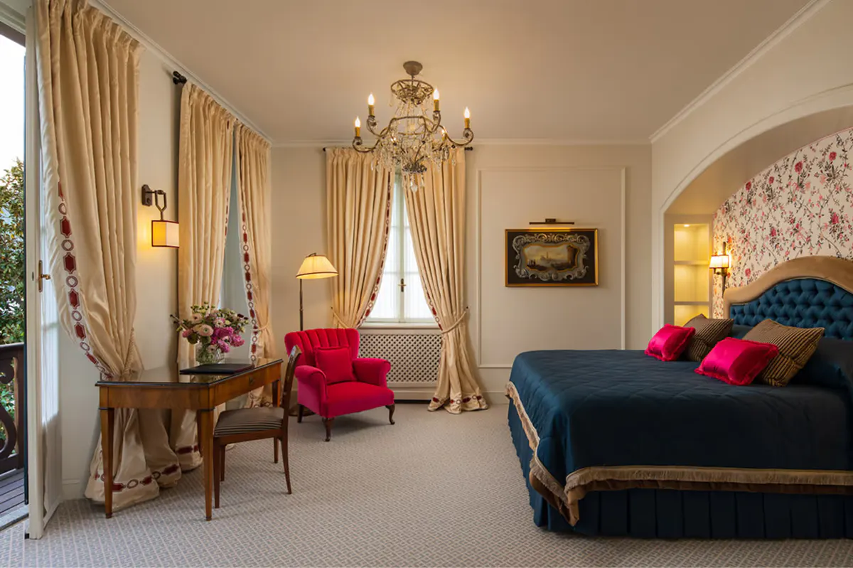 Suite bedroom with red velvet seating, gold bed frame, frescoed ceiling, and arched window
