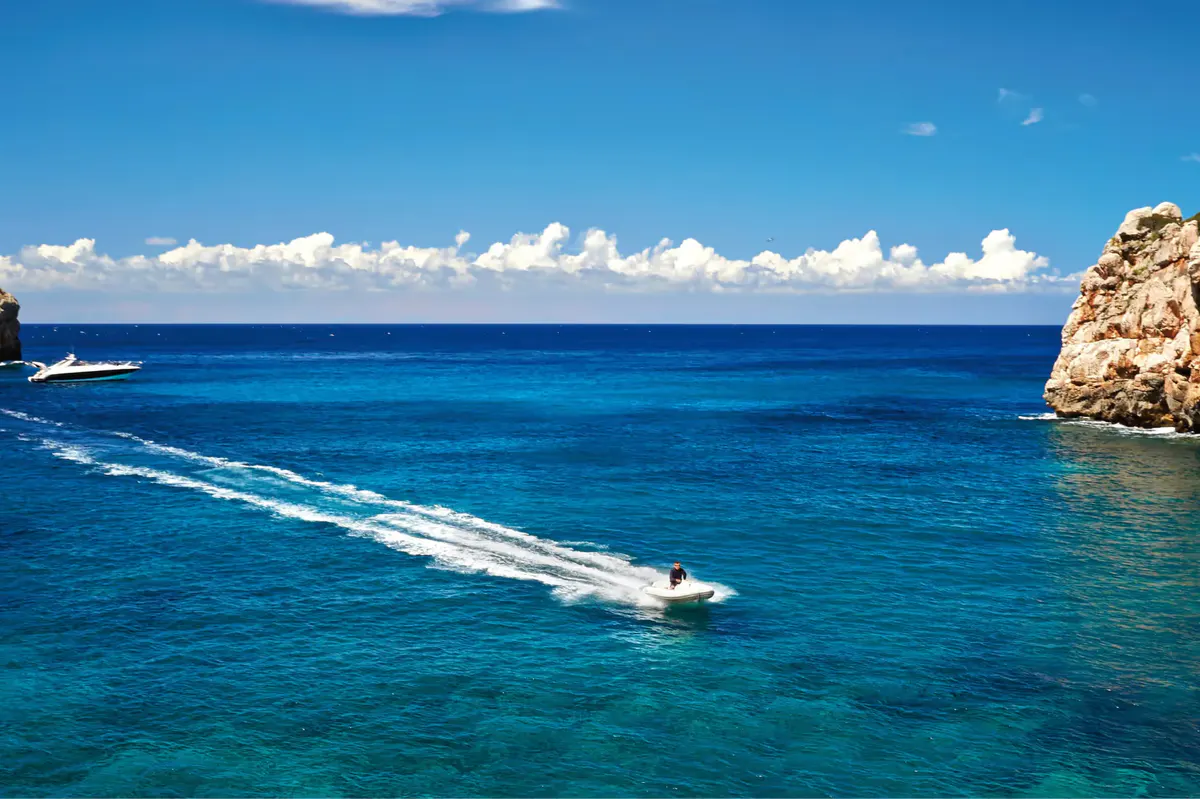 Boat traveling on turquoise Mediterranean water with rocky coastline and clear sky