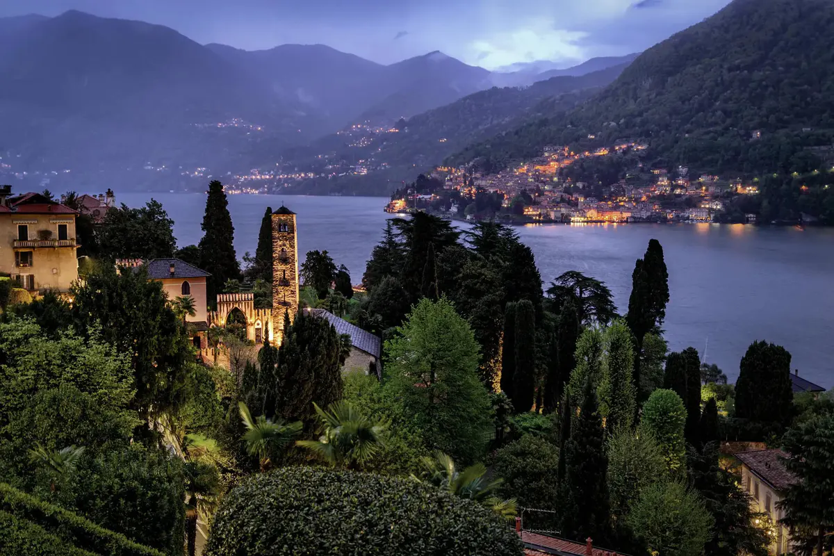 Twilight view of lit palazzo and cypress with lake and town lights below