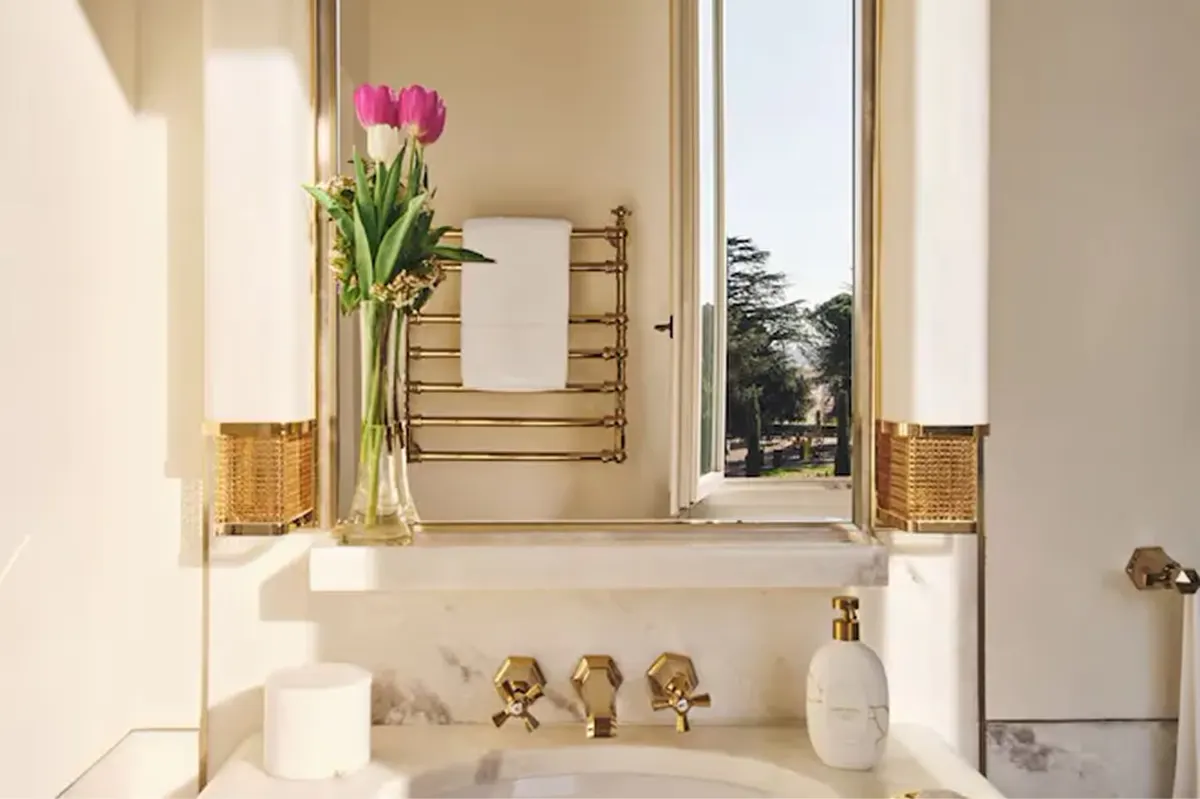 A marble bathroom with a vanity, brass fixtures, fresh flowers in a vase, and natural light from a window