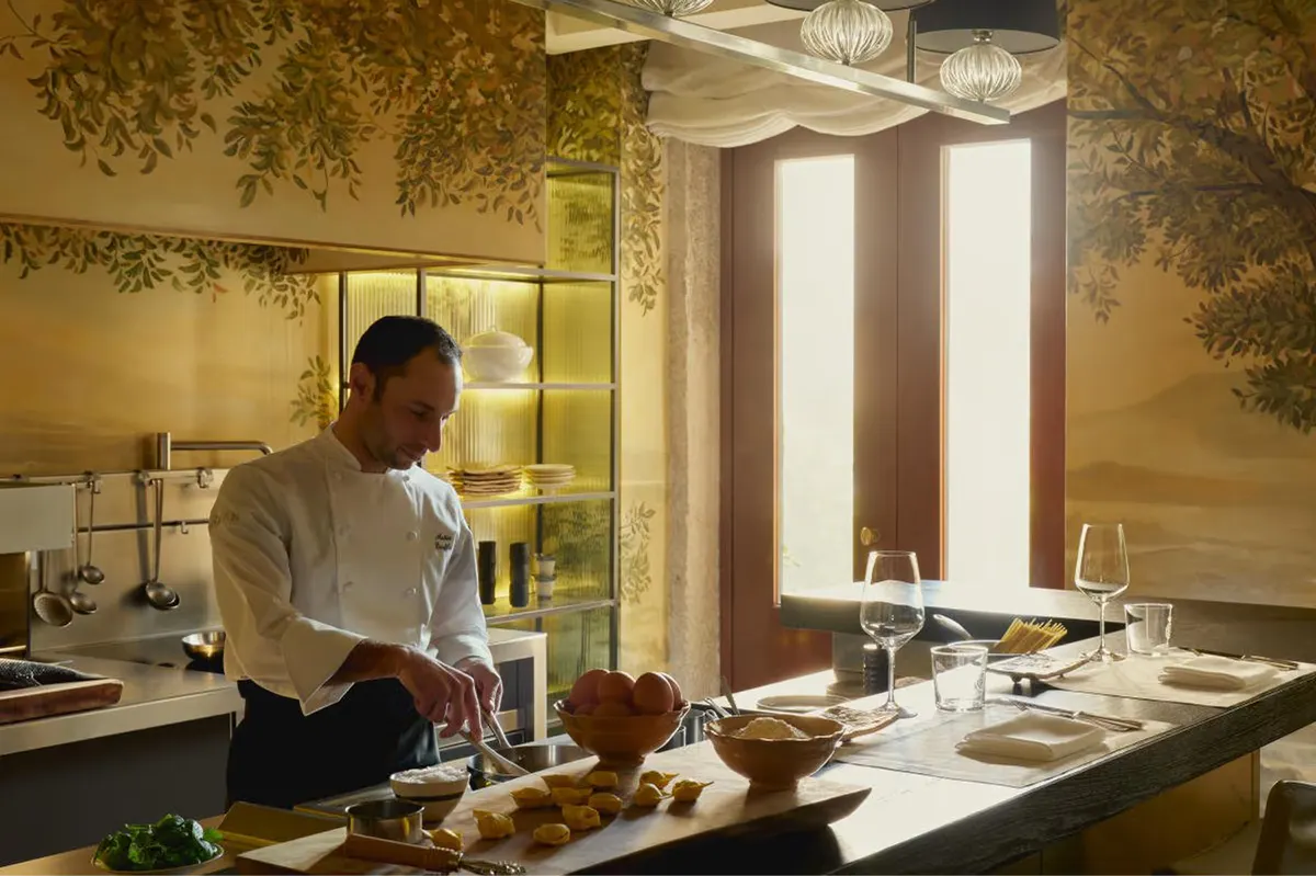 An open kitchen with a chef preparing pasta, warm lighting overhead, and ceramic bowls of ingredients nearby