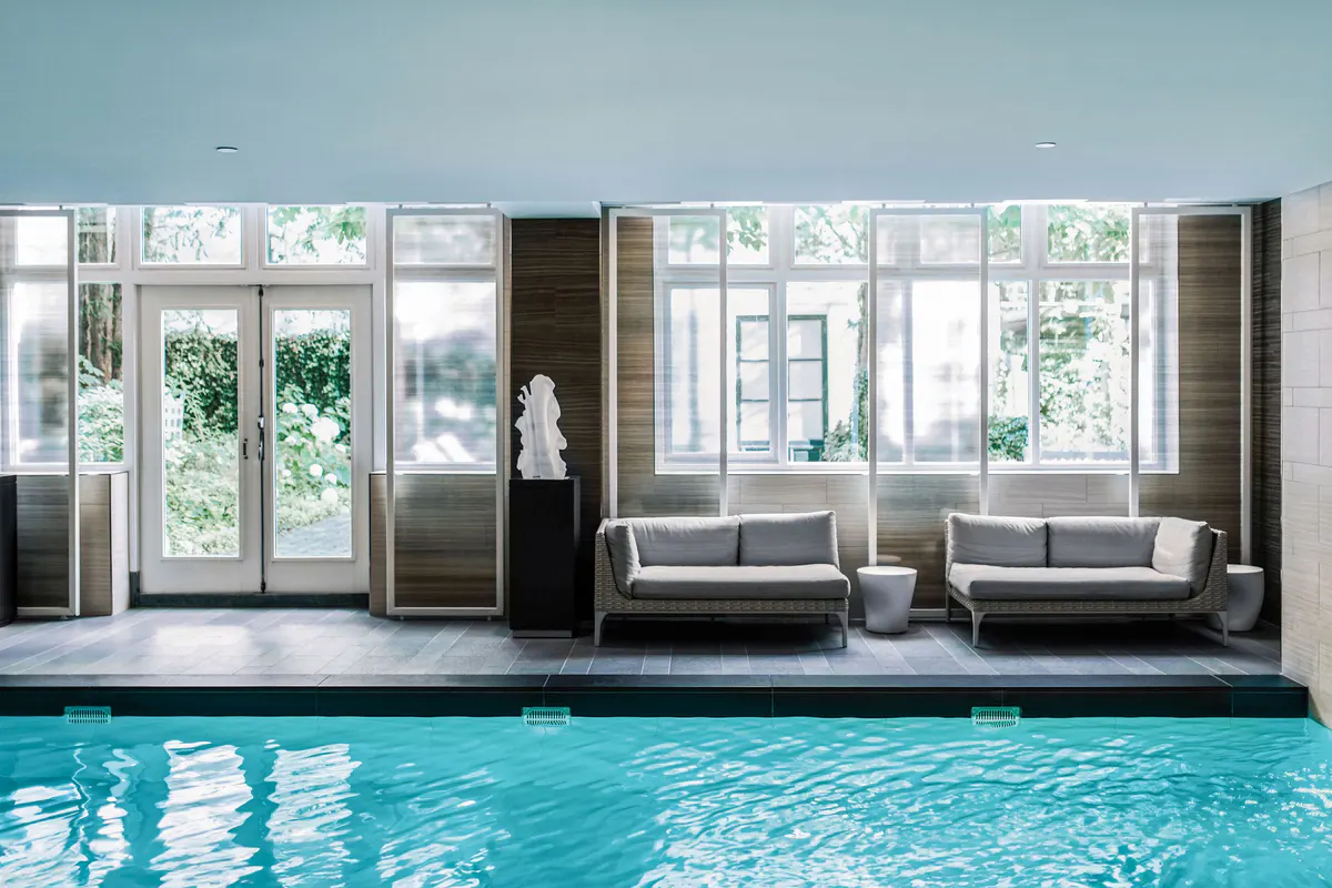 An indoor pool with bright turquoise water flanked by lounge seating and tall windows framed in dark wood