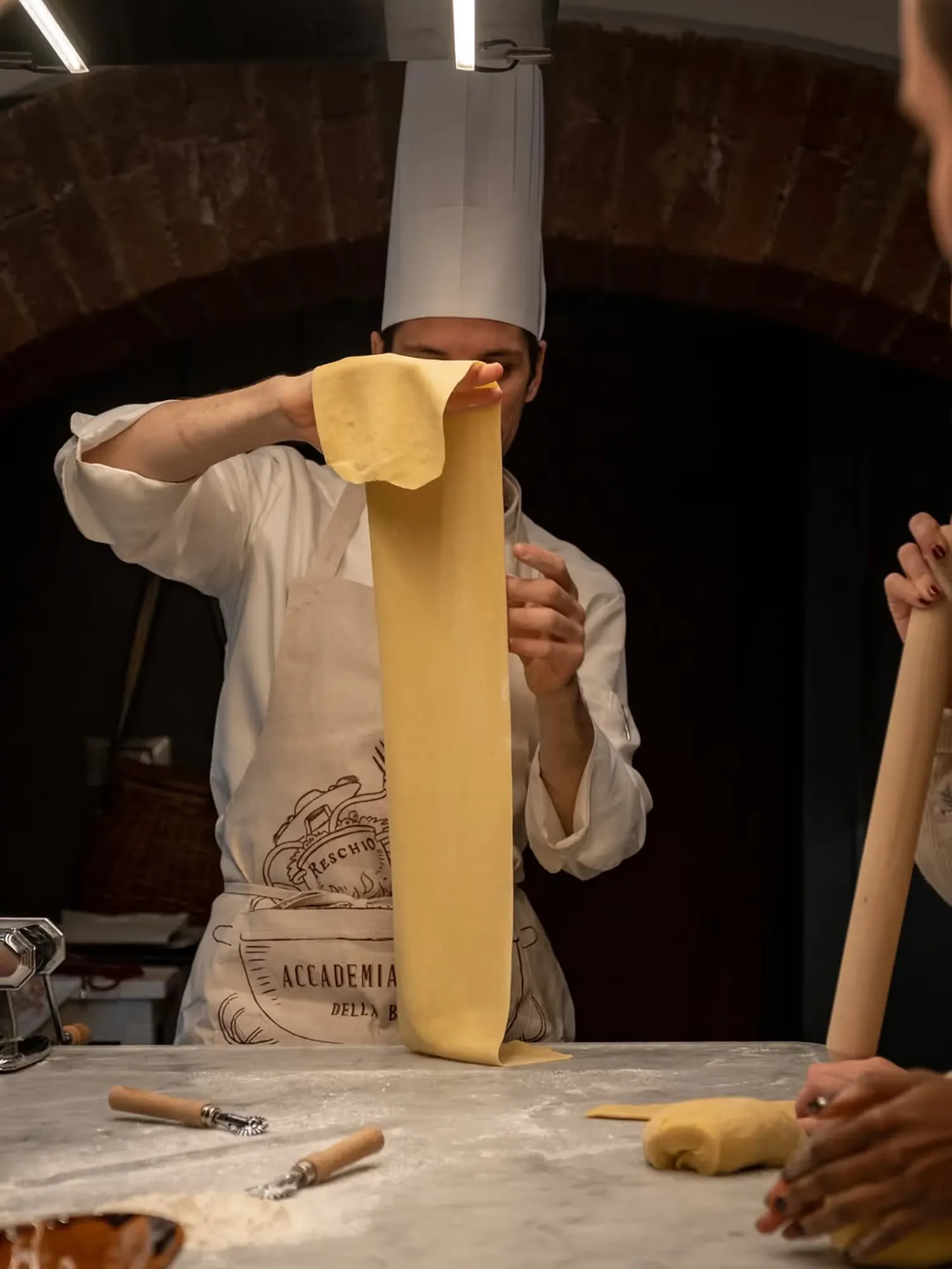 A chef stretching fresh pasta by hand in a vaulted kitchen