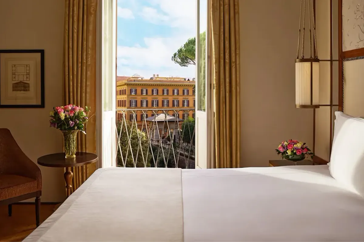 A bedroom window framing a view of a Rome palazzo’s dome and roofscape, with white bedding in the foreground