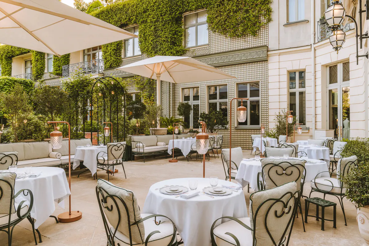 An outdoor terrace dining area with white tablecloths, bentwood chairs, and garden surroundings