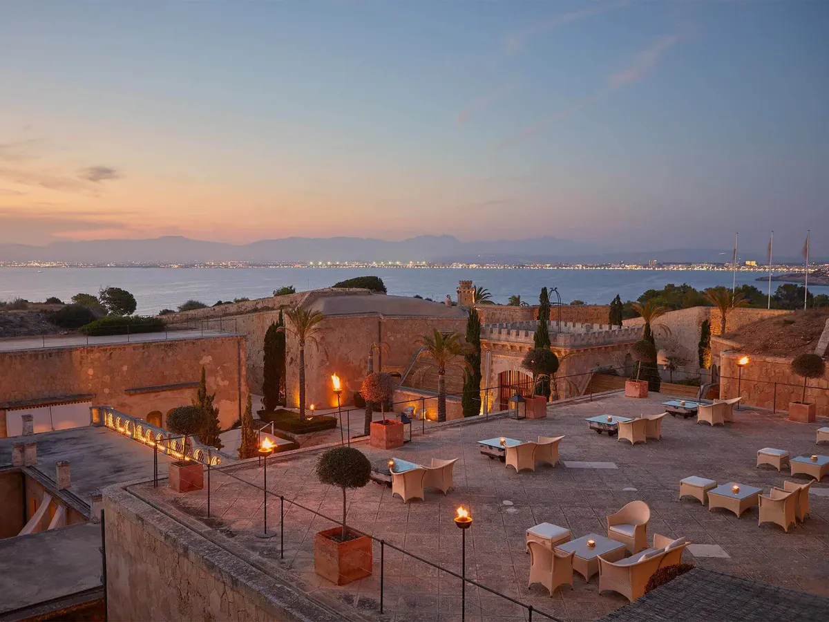 A rooftop terrace at twilight with cream loungers, lit lanterns, and sea horizon