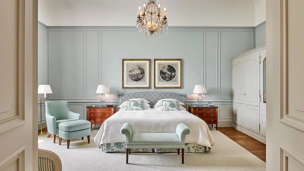 A guest bedroom with soft green walls, a white bed, matching armchairs, and a crystal chandelier