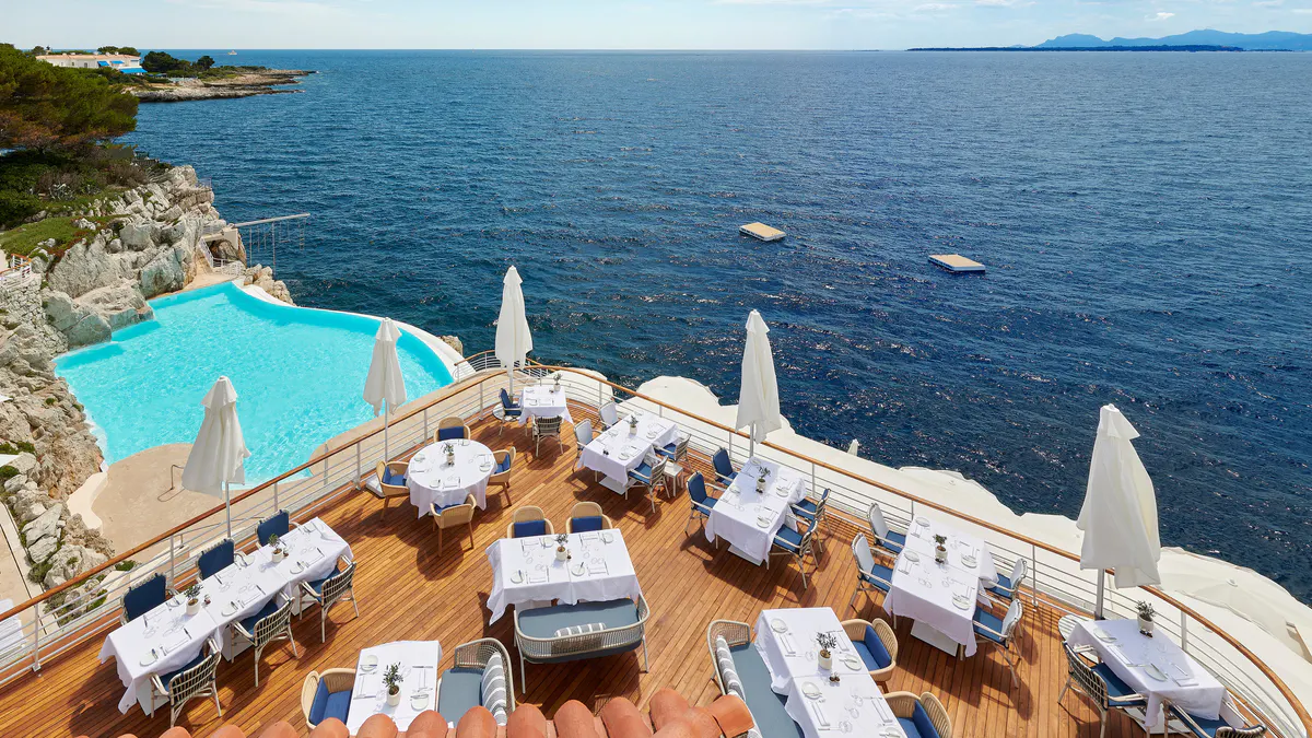 An open-air terrace restaurant with white umbrellas, sea views, and tables overlooking the Mediterranean