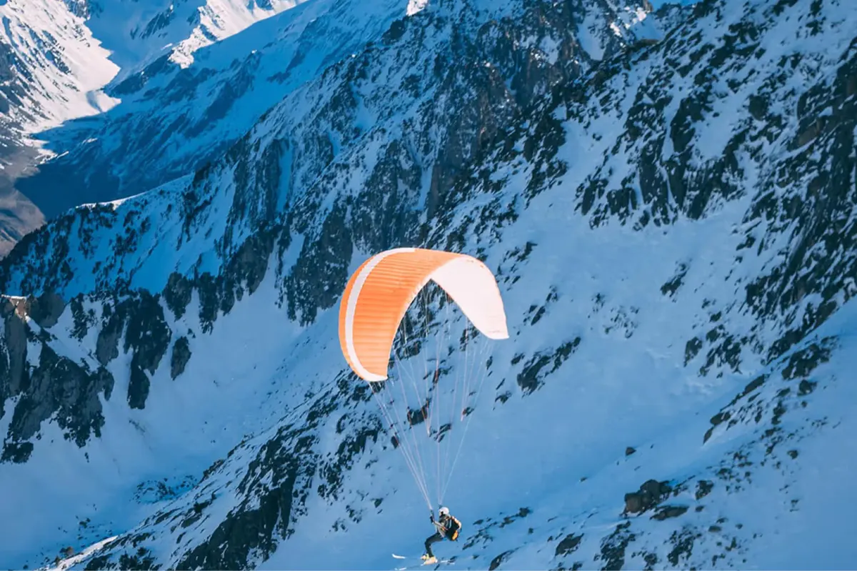 Paraglider in orange parachute soaring above snow-covered alpine mountains