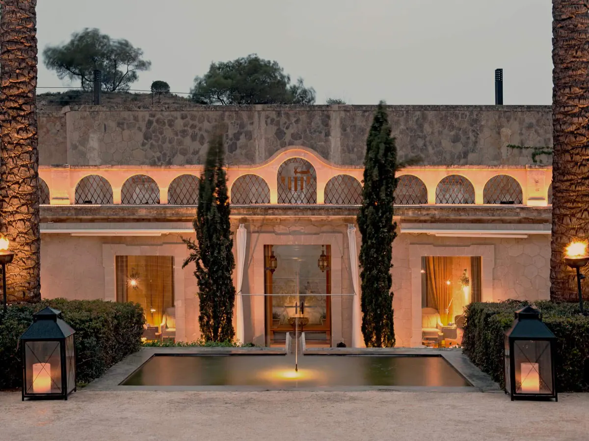 A cream and terracotta fortress facade with rounded arched windows, flanked by tall cypress trees and lanterns
