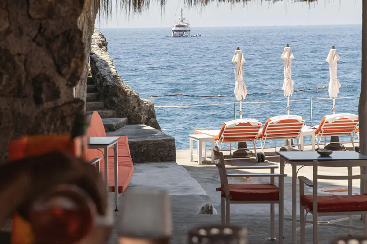 Private cove with orange striped loungers, white umbrellas, rocky cliff overhang, calm blue water and distant boat