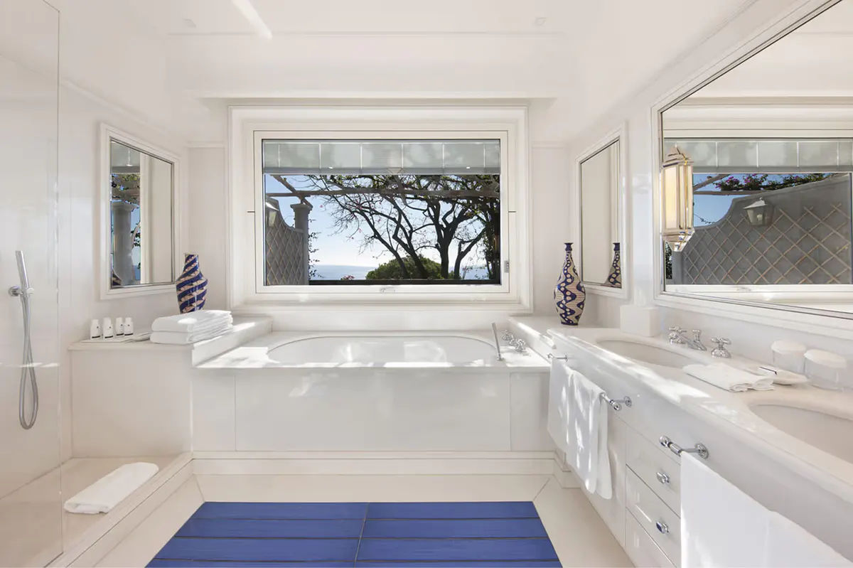 White marble bathroom with large window framing rocky coastline, blue accent mat below freestanding soaking tub