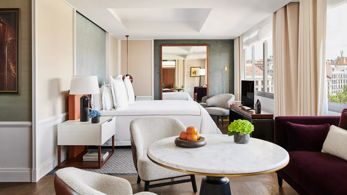 A suite with white bedding, sitting area, and glass partition separating the living space