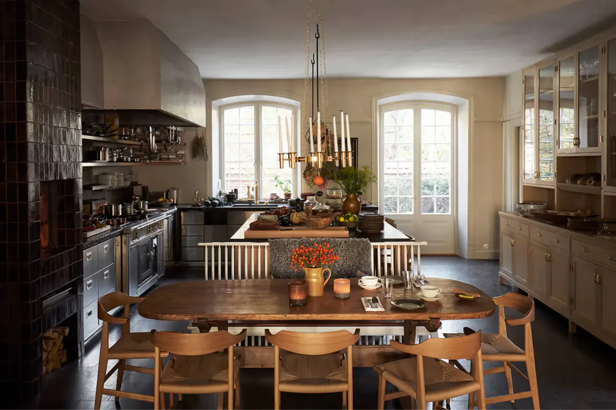 A kitchen dining room with wooden table, leather chairs, open shelving, and two tall arched windows
