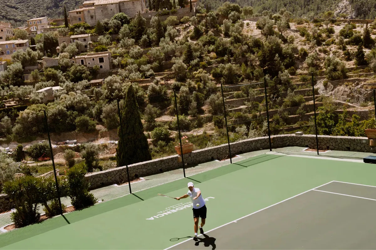 Tennis court on hillside with player, surrounded by Mediterranean vegetation and cypress trees